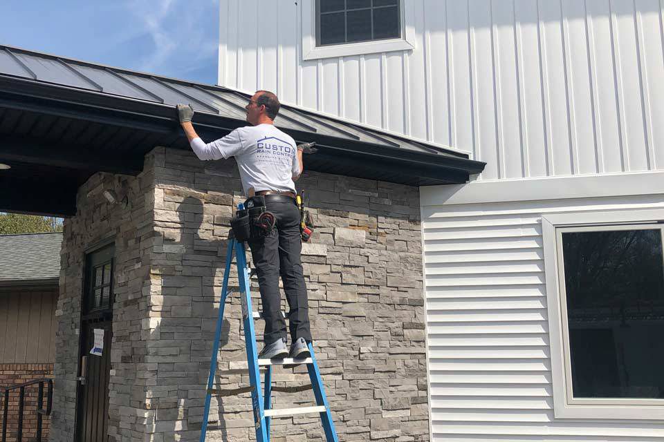 Custom Rain Control employee wearing a uniform shirt with logo installing custom rain gutters on the side of a house