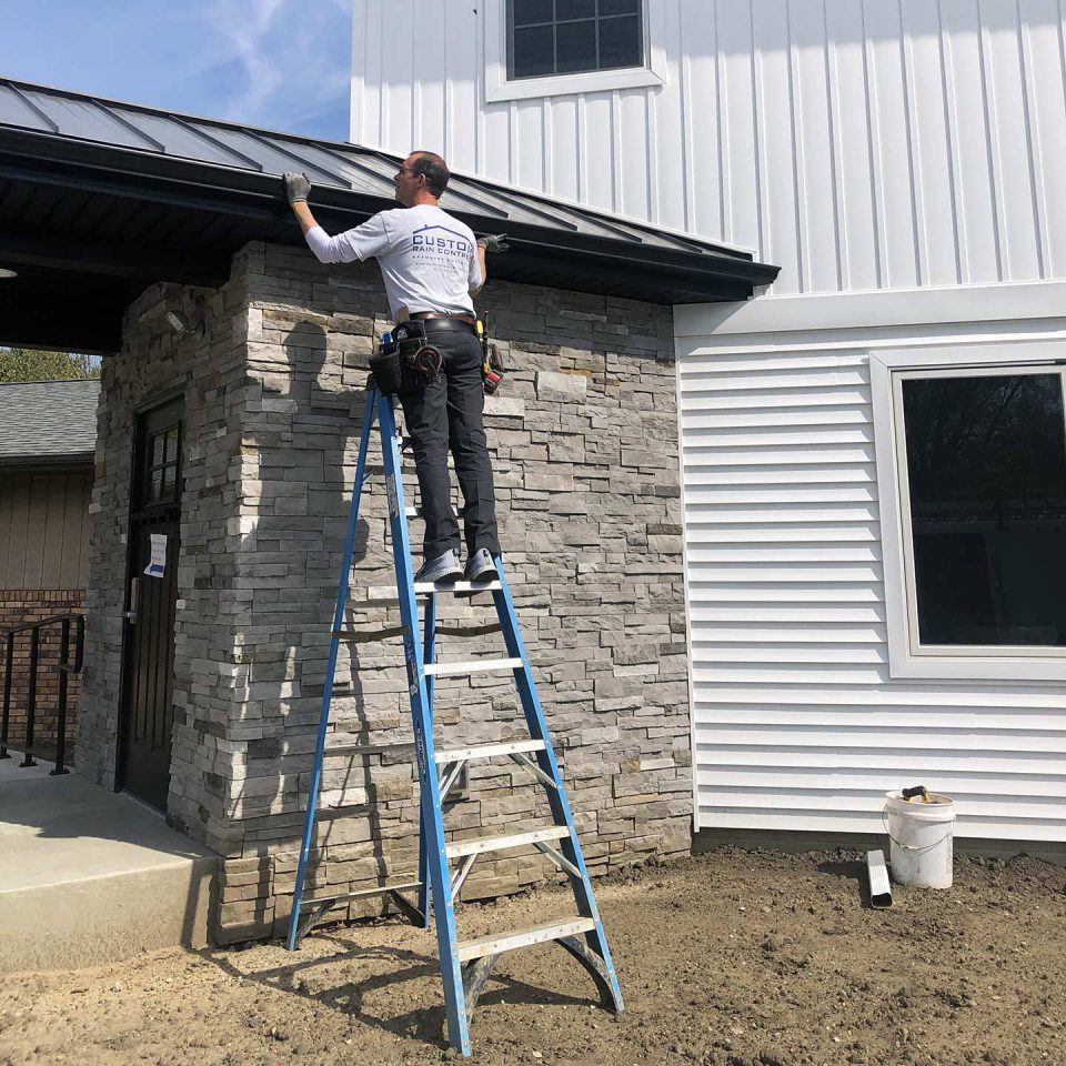 Custom Rain Control employee wearing a uniform shirt, standing on a ladder to install a rain gutter system