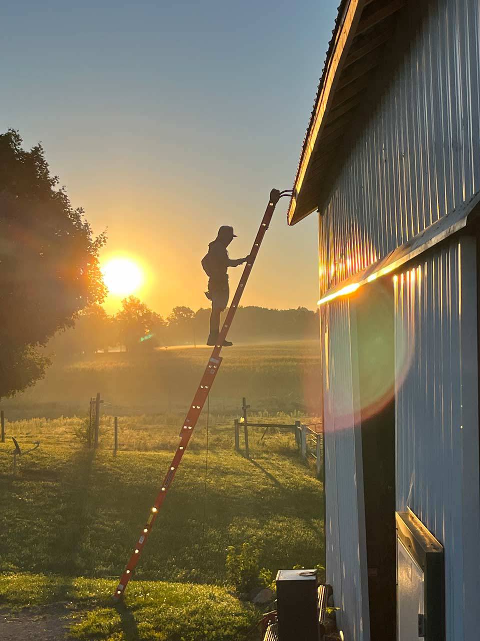 Man standing on a tall ladder after installing a gutter system on the roof of a barn with the sun behind him